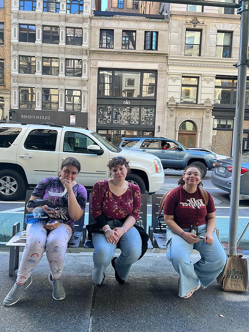 3 students sitting on bench with Fifth Avenue behind them