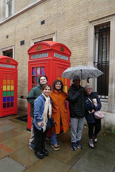 在伦敦的一条街上，五个学生站在一个鲜红色的老式电话亭前. 学生们都穿着雨具，其中一个还撑着伞