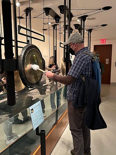 Man wearing baseball cap and protective face mask hitting a gong with a mallet inside museum