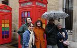 在伦敦的一条街上，五个学生站在一个鲜红色的老式电话亭前. 学生们都穿着雨具，其中一个还撑着伞